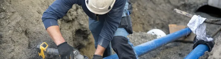 Un ouvrier portant un casque et des gants coupe un tuyau d'eau bleu dans une tranchée.
