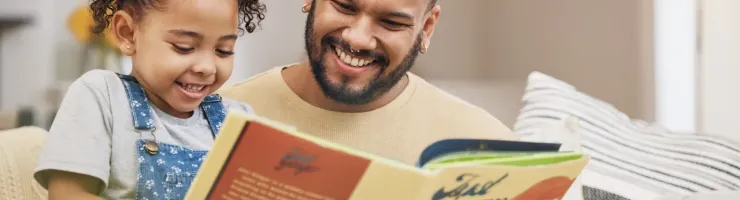 Père et fille souriants lisent ensemble un livre d'histoires sur le canapé. Moment de complicité et d'éducation.