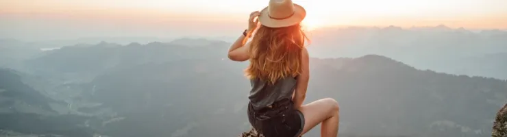 Une femme de dos, portant un chapeau, est assise sur un rocher et contemple un panorama montagneux au coucher du soleil.