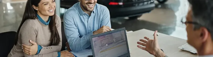 Dans une concession automobile, un couple consulte un vendeur à une table avec un ordinateur portable pour acheter un véhicule.