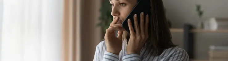 Une femme parle au téléphone, debout devant une fenêtre, avec un éclairage naturel.