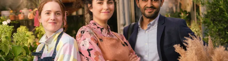 Deux femmes en tablier, à côté d’un homme en costume, sont devant une boutique.