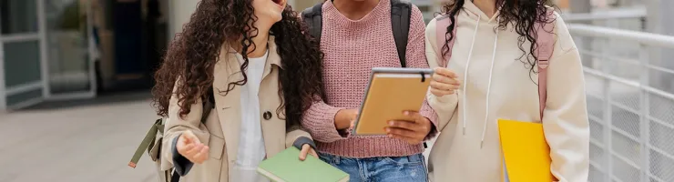 Trois jeunes étudiantes marchent ensemble, portant des livres et des dossiers sous le bras, souriant et discutant.