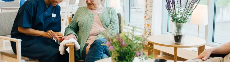 Une infirmière assise à côté d'une femme âgée dans un fauteuil, échangeant des sourires et des paroles réconfortantes.