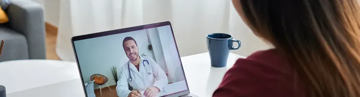 Une femme assise à un bureau, utilisant un ordinateur portable avec un médecin visible sur l'écran.