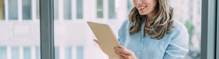 Portrait d'une femme souriante en chemise bleue, debout devant une grande fenêtre de bureau, regardant et tenant une tablette numérique.