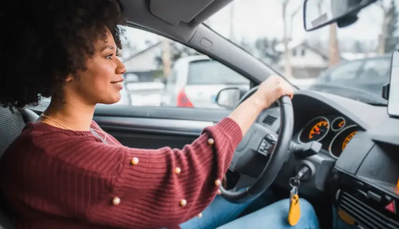 Une femme conduit une voiture en souriant, vue depuis l'intérieur du véhicule.