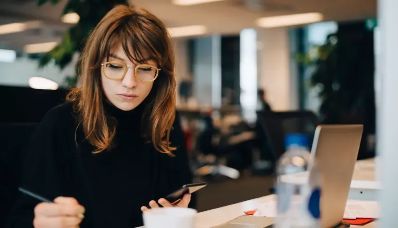 Une femme est en train de consulter son téléphone portable et écrire quelques chose sur une feuille. 