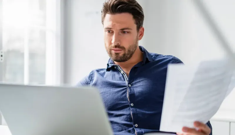 Un homme consulte des documents devant son ordinateur portable, concentré sur ses tâches administratives.