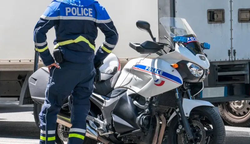 Un policier en uniforme se tient près de sa moto, prêt à intervenir sur la route.