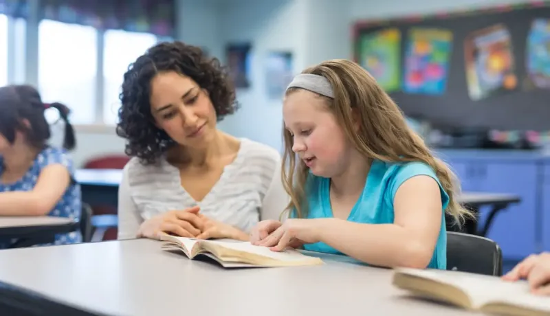 Une femme aide une jeune fille à faire ses devoirs dans une salle de classe lumineuse.
