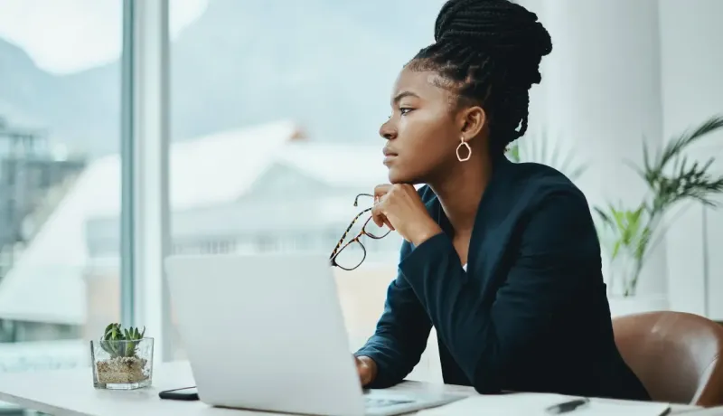 Femme pensive regardant par la fenêtre avec un ordinateur devant elle.
