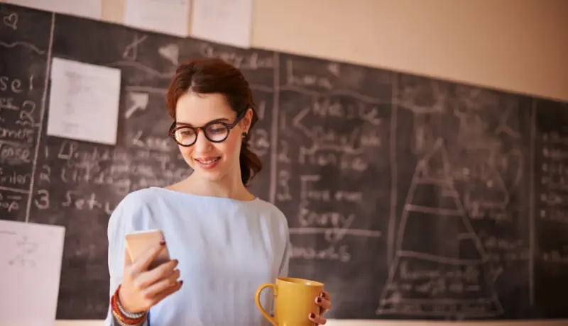 Femme souriante regarde son téléphone en tenant une tasse jaune devant un tableau noir.