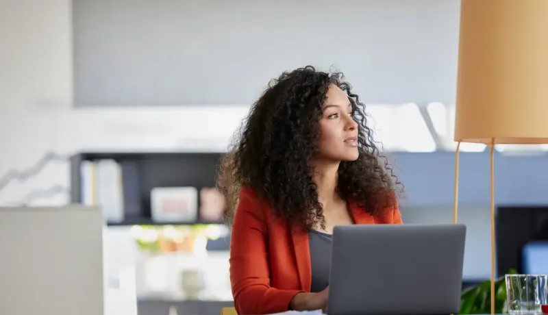 Femme aux cheveux bouclés assise à un bureau avec un ordinateur portable, regardant pensivement sur le côté.