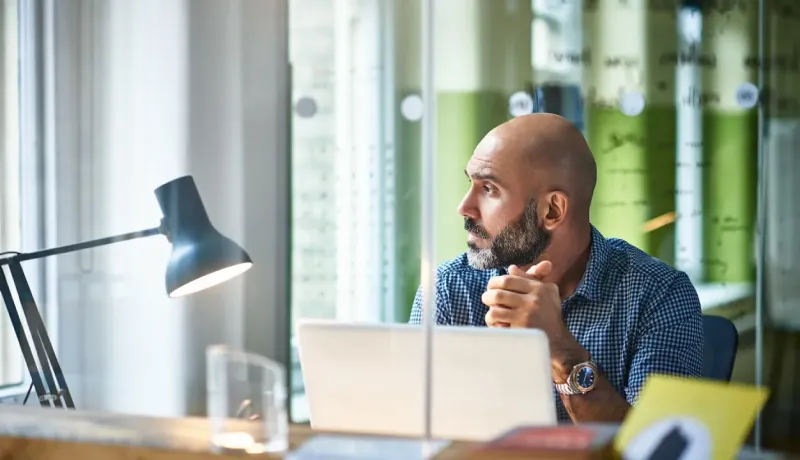 Homme chauve avec barbe assis à un bureau avec un ordinateur portable, regardant sur le côté.