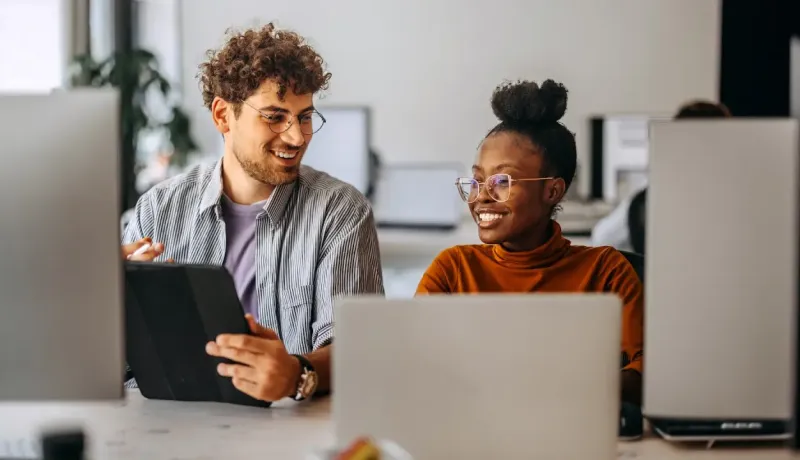 Deux collègues souriants regardent un écran de tablette dans un bureau.