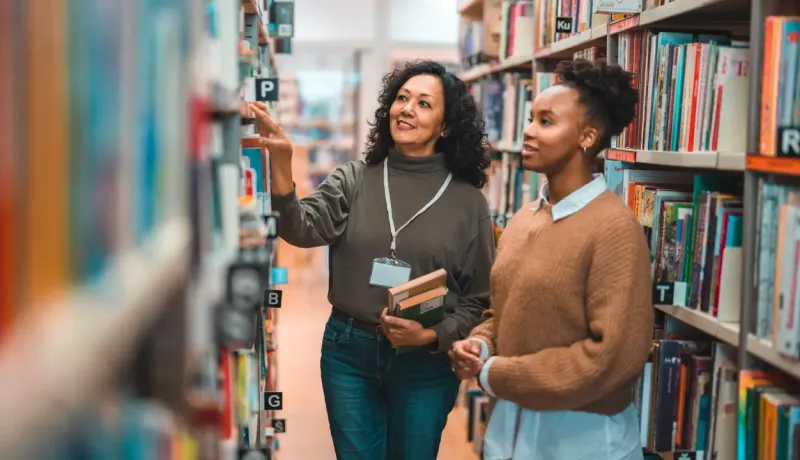 Une bibliothécaire et une étudiante discutent ensemble dans une bibliothèque.