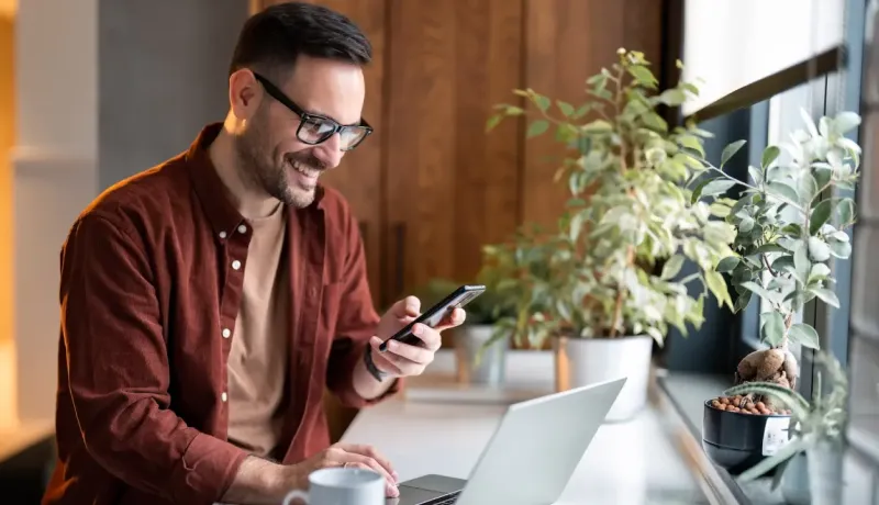 Homme souriant avec lunettes, utilisant téléphone et ordinateur portable près d'une fenêtre avec plantes.