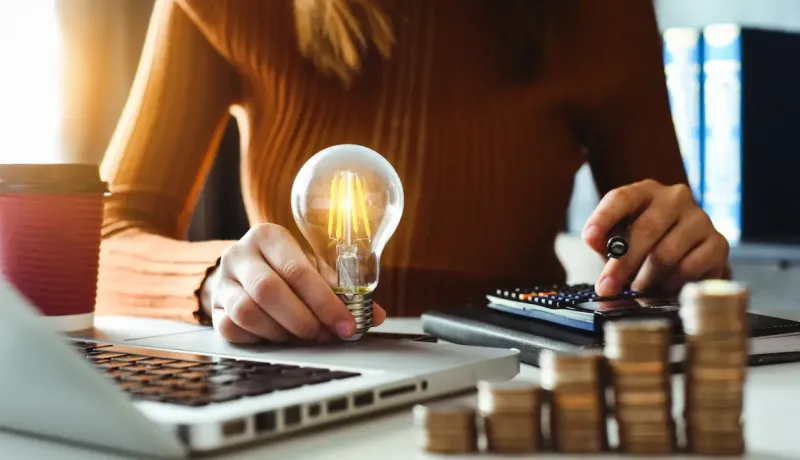 Personne tenant une ampoule et utilisant une calculatrice près d'un ordinateur portable et de piles de pièces.