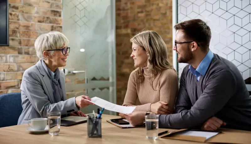 Un conseillère bancaire est en train de conseiller un couple. Elle tend un document au couple dans sa main.