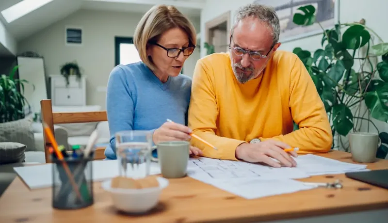 Un couple âgé examine des papiers financiers à table, discutant de leur gestion budgétaire.