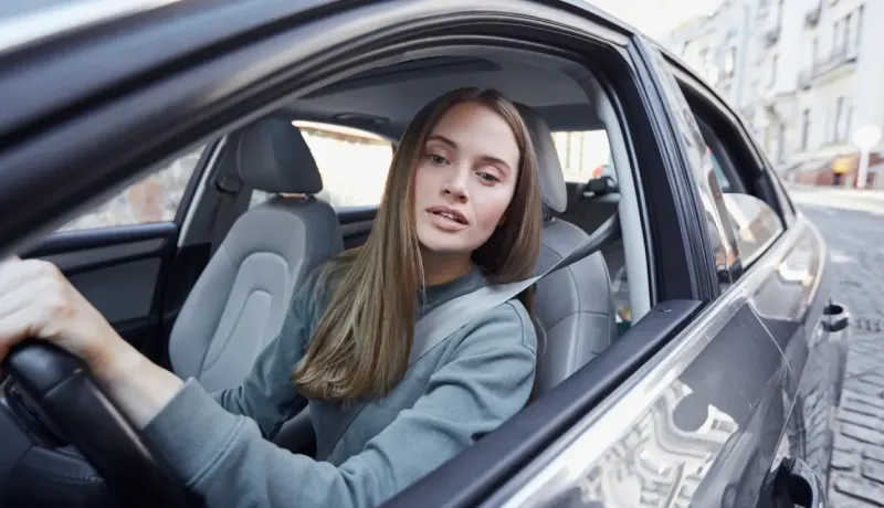 Femme aux longs cheveux blonds assise au volant d'une voiture, regardant par la fenêtre.