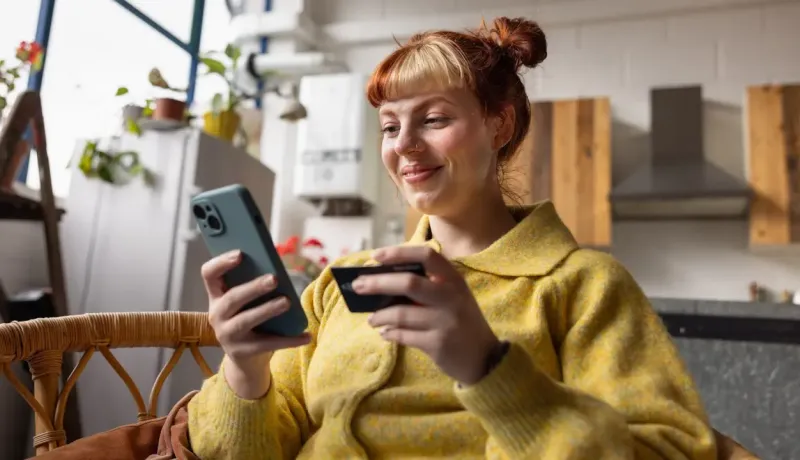 Femme souriante assise dans une chaise, tenant un téléphone et une carte bancaire.