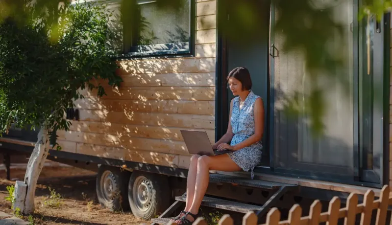 Femme assise sur les marches d'une tiny house en bois, sur un ordinateur portable.