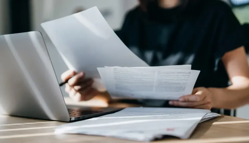 Personne assise à un bureau avec un ordinateur portable, examinant des documents.