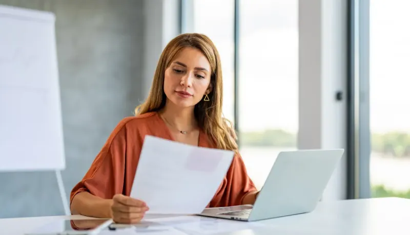 Une femme regarde une feuille et travaille avec un ordinateur portable.