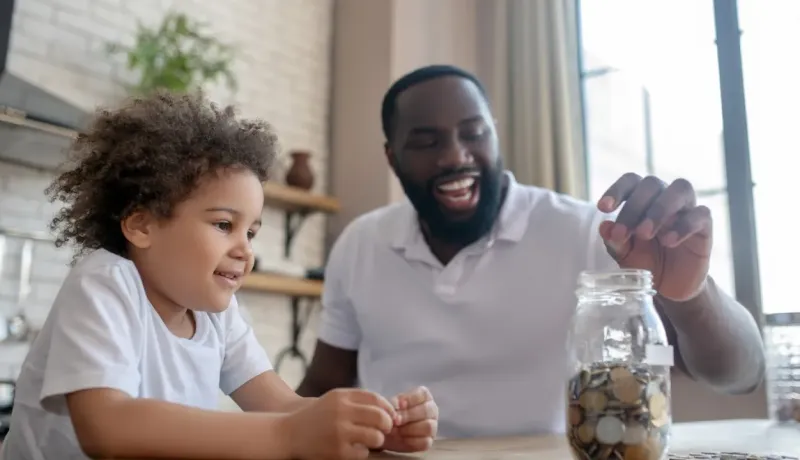 Un papa souriant met une pièce dans un bocal et montre à son fils comment économiser.