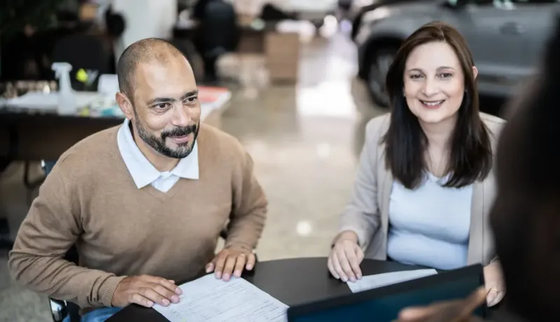 Couple assis à une table avec un vendeur, examinant des documents dans une concession automobile.
