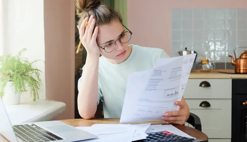 Jeune femme dépassée consulte des documents financiers dans sa cuisine.