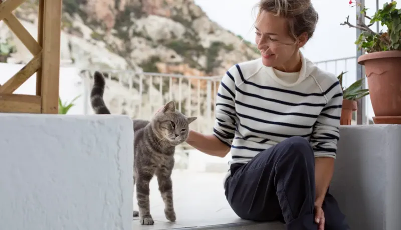 Une femme en pull rayé caresse un chat tigré gris sur un patio extérieur ensoleillé.