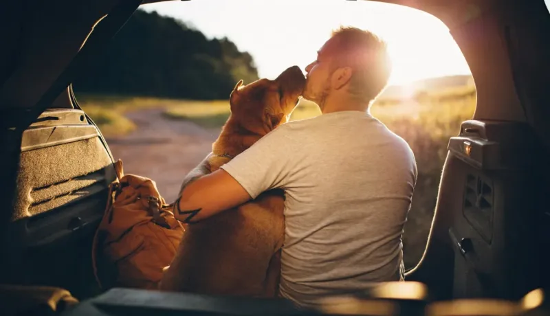 Un homme embrasse un chien de profil, assis à l'arrière d'une voiture avec le coffre ouvert.