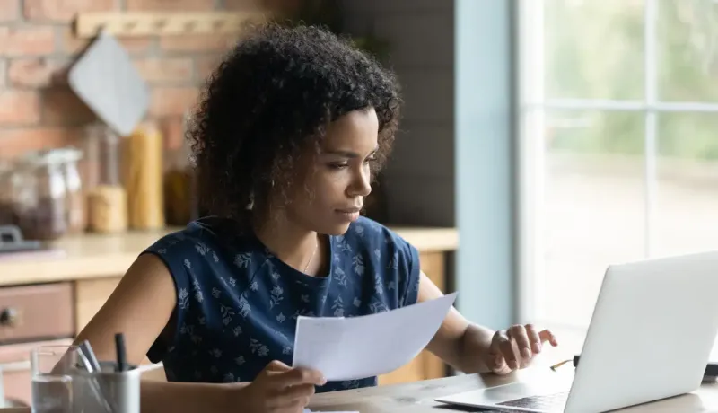 Une femme, tenant un document, utilise son ordinateur portable dans un bureau à domicile.