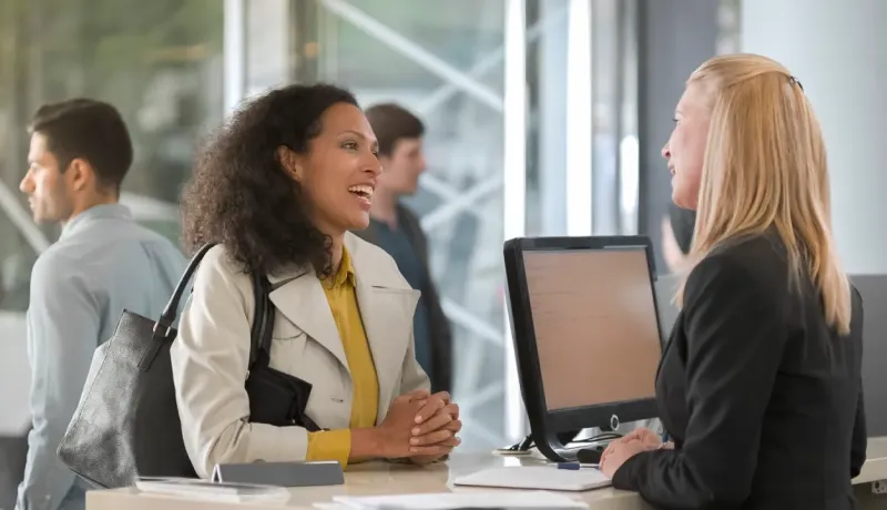 Une femme souriante parle à une employée derrière un comptoir, possiblement à une banque ou réception.