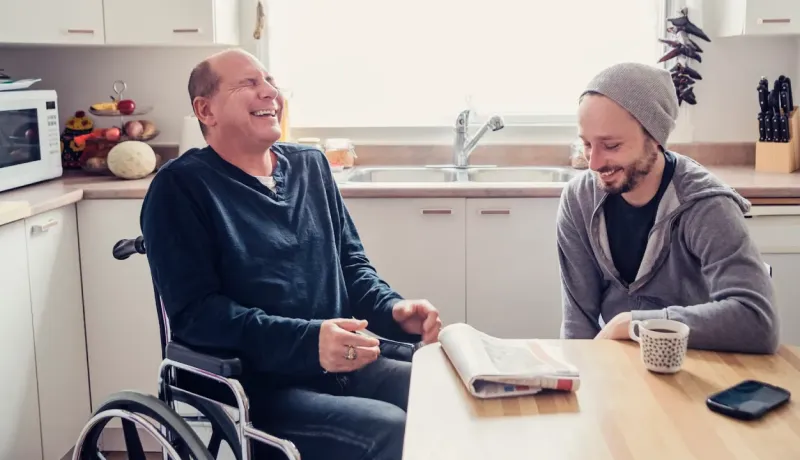 Deux hommes rient ensemble dans une cuisine, l'un en fauteuil roulant et l'autre assis à une table.