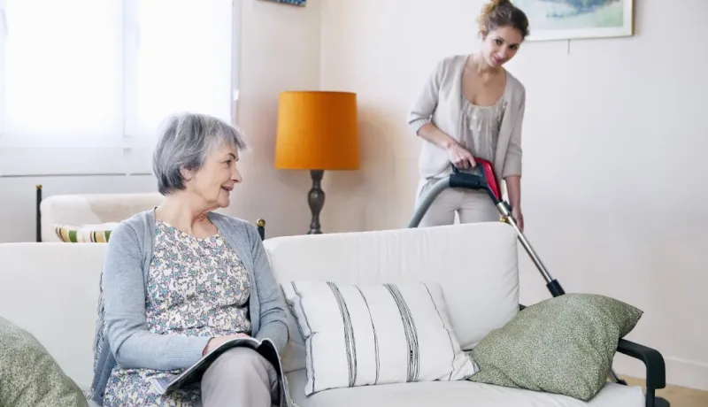 Une jeune femme passe l'aspirateur pendant qu'une femme âgée est assise sur un canapé.