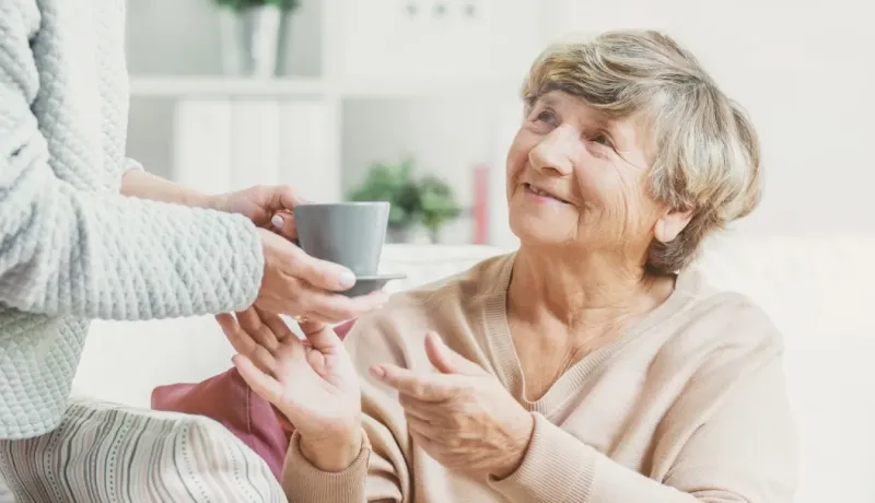 Une main offre une tasse de thé ou café à une femme âgée souriante.