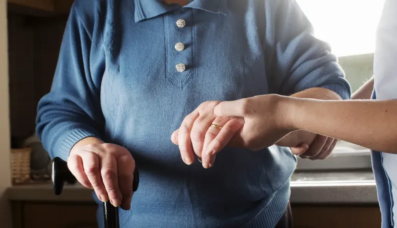 Gros plan des mains d'une aide soignante tenant la main d'une personne âgée s'appuyant sur une canne.