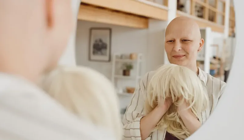 Une femme chauve tenant une perruque blonde se regarde dans un miroir, souriante.