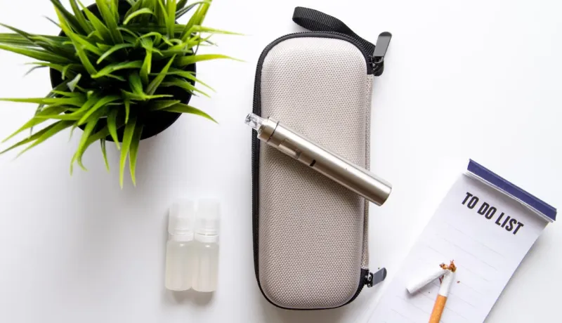 Une cigarette électronique est posée sur une coque de protection, des recharges pour la cigarette électronique se trouve à gauuche de l'étui de protection. Une cigarette cassée est sur une liste de choses à faire.