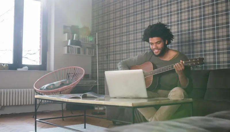 Un homme souriant aux cheveux bouclés joue de la guitare acoustique en regardant un ordinateur portable.