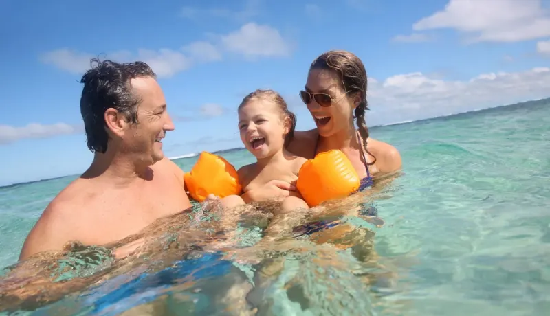 Une famille joyeuse : père, mère et leur fille s'amusent dans l'eau turquoise sous un ciel bleu.