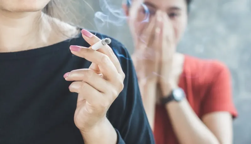 Une personne fume une cigarette, la fumée s'échappant, tandis qu'une autre se couvre le nez en arrière-plan.