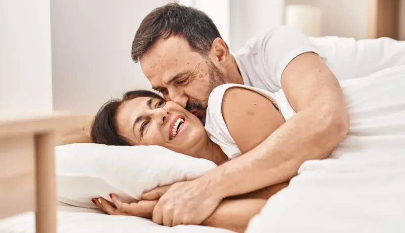 Un couple sourit et s'enlace tendrement au lit, l'homme embrassant la joue de la femme.