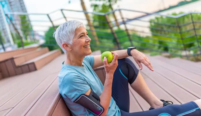 Une femme aux cheveux gris, assise à l'extérieur, sourit en tenant une pomme verte après le sport.