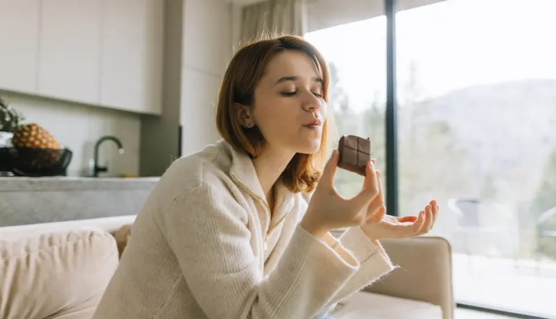 Une jeune femme aux cheveux roux savoure un morceau de chocolat, les yeux fermés.