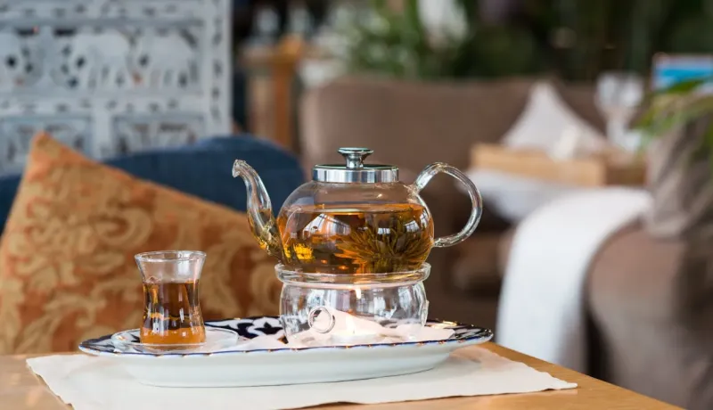 Une théière en verre avec du thé fleuri est posée sur un chauffe-thé, à côté d'un petit verre.